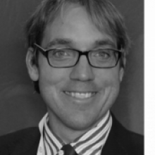 Black and white headshot of Professor Davide Forcellini with black rimmed glasses, striped button up and blazer.