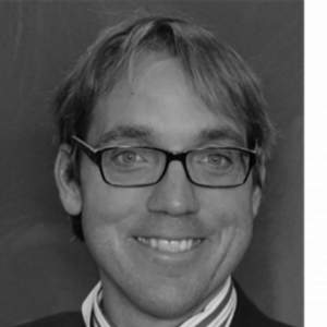Black and white headshot of Professor Davide Forcellini with black rimmed glasses, striped button up and blazer.
